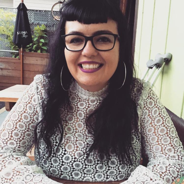 Alisha with long black hair and Betty bangs, wearing glasses, hoop earrings, red lipstick, and a white lace shirt. Her crutches are behind her.