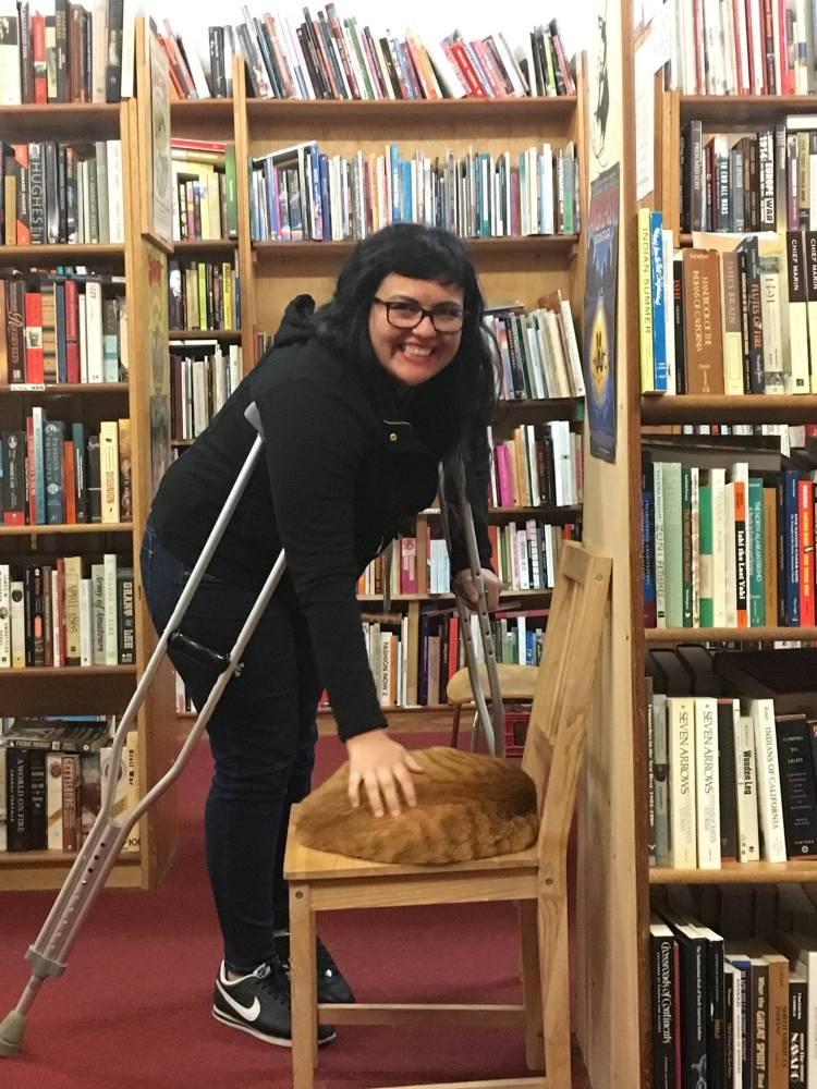 Alisha wearing all black, leanng on her crutches and petting an orange cat inside a bookstore.