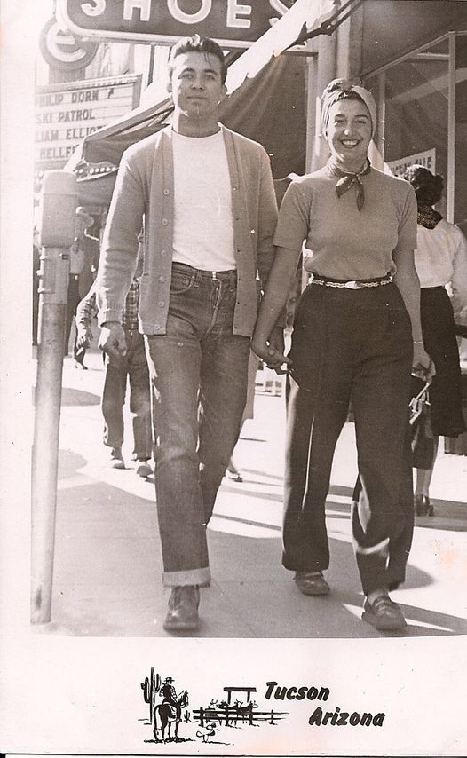 a black and white photo of Louis Vasquez and Theadora Petras holding hands, smiling, and walking down Congress Street in 1944. Louis is on the left, wearing a white t-shirt, a cardigan, and Levi’s. Theadora is on the right, wearing a shirt, pants with a fancy belt, and her hair pulled back in a headscarf.
