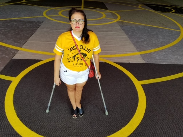 Alisha, a woman with medium-light skin and black hair, stands with crutches in the middle of a large painted yellow circular design on the floor. She is wearing coral lipstick, black rimmed glasses, and looks away from the camera. Her hair falls over her left shoulder, over a yellow baseball tee with black text featuring a cartoon image of Dock Ellis. She is wearing white shorts and black sandals, and has a red purse slung across her body.