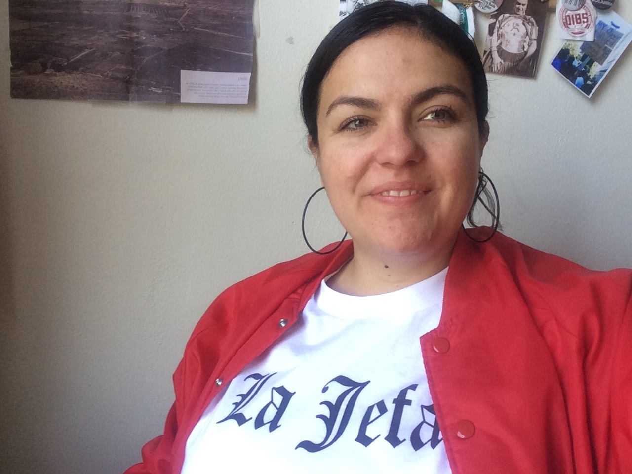 Alisha wearing a red jacket, white shirt with the words, "La Jefa" in old english, and black hoop earrings with her dark hair pulled back.